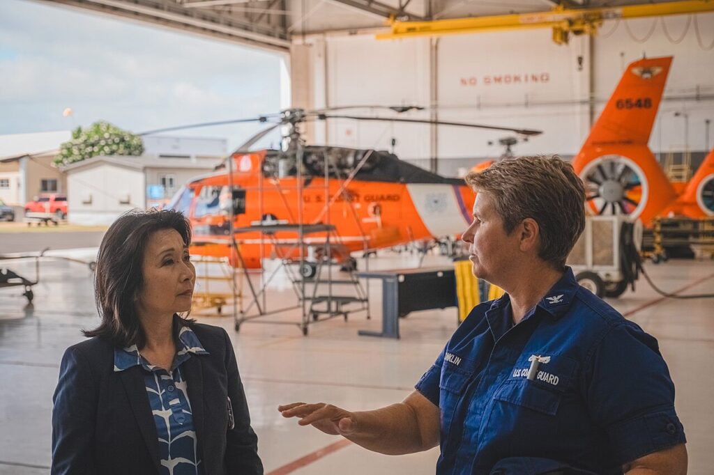 Two professionals in a hangar talk beside an orange Coast Guard helicopter on the tarmac around maintenance equipment.