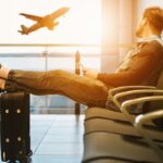 Man relaxing in an airport terminal with feet on a rolling suitcase, watching a plane take off outside through the window.