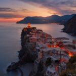 Sunset over a cliffside Italian harbor town with glowing lights and pastel buildings along the coast, calm sea to the left.