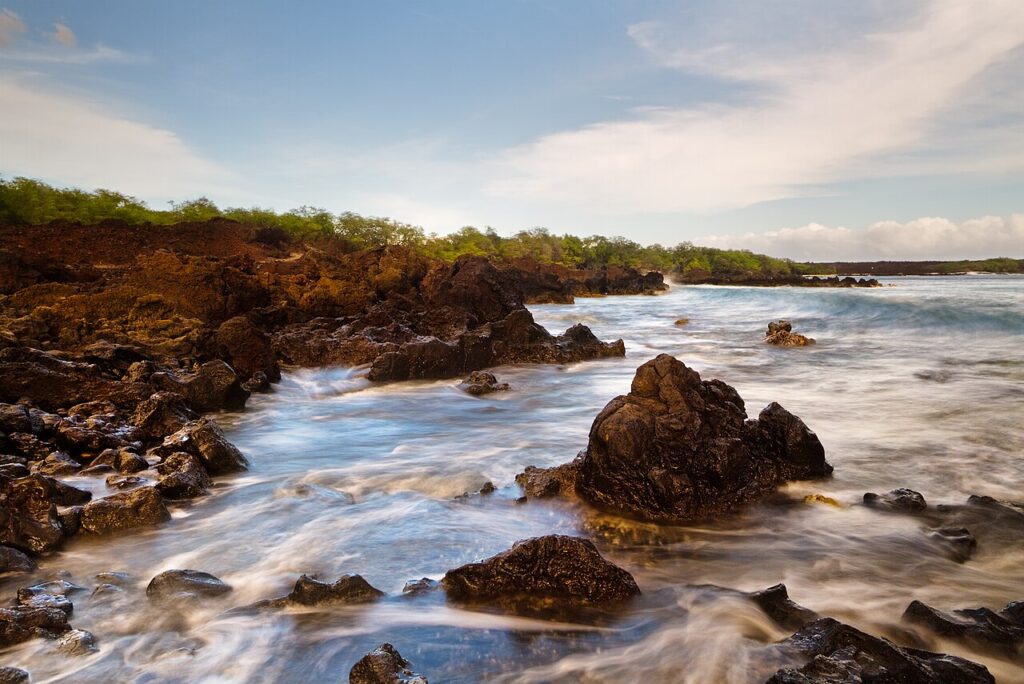 Rugged coastal rocks along a calm shoreline with misty waves washing over dark lava rocks under a blue sky with wispy clouds (long exposure)