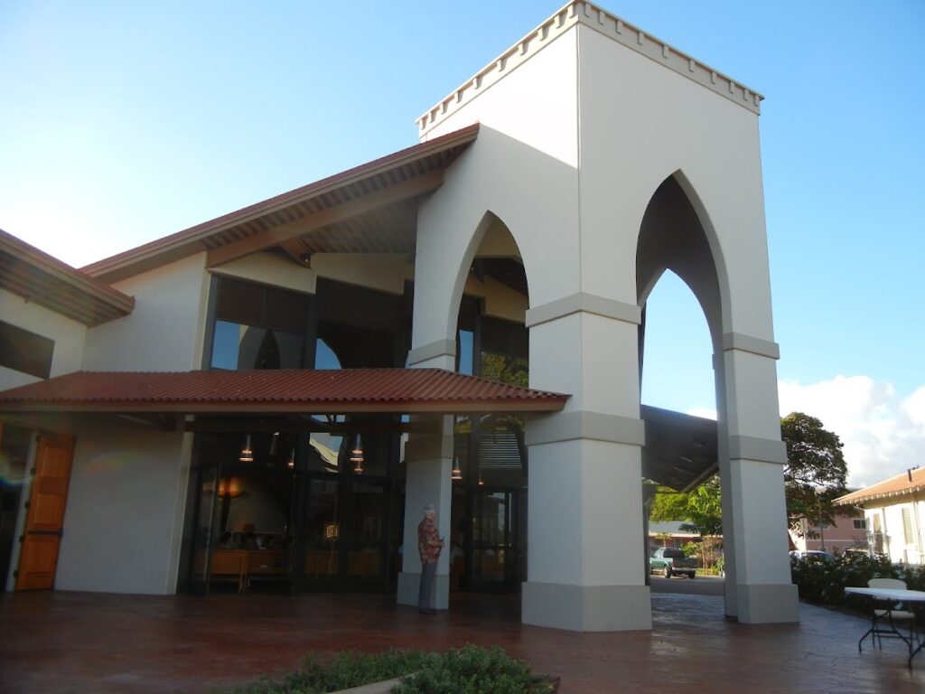 Modern building with tall arched openings and a red-tiled roof, glass entry doors, and a clear blue sky above. A person stands near the entrance.