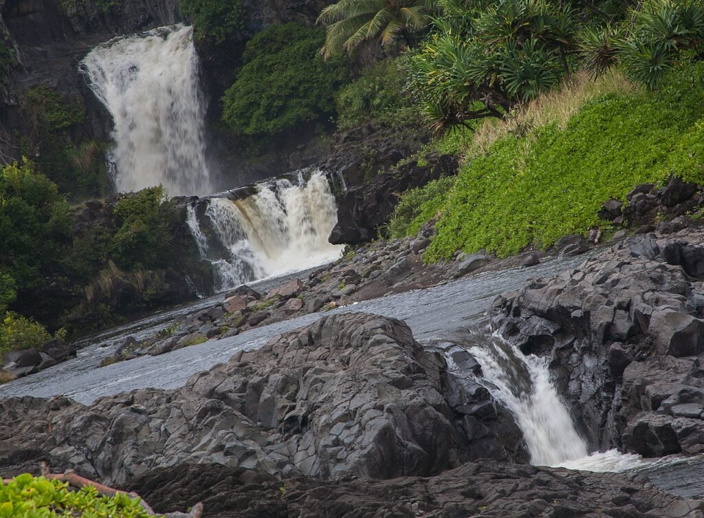 Travel Guide Highlights Seven Hawaii Long Weekend Destinations - Maui Tom's Vacation Rental Cleaning & Maintenance Waterfall cascading down dark rocks into a rocky river with surrounding green vegetation and trees nearby, tropical scene.