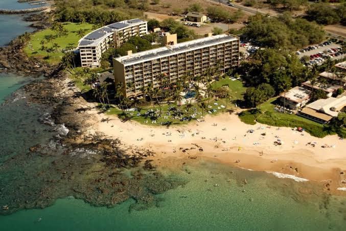 Aerial view of a beachfront resort with a multi-story hotel surrounded by palm trees, adjacent sandy beach, and turquoise water along a rocky shoreline.