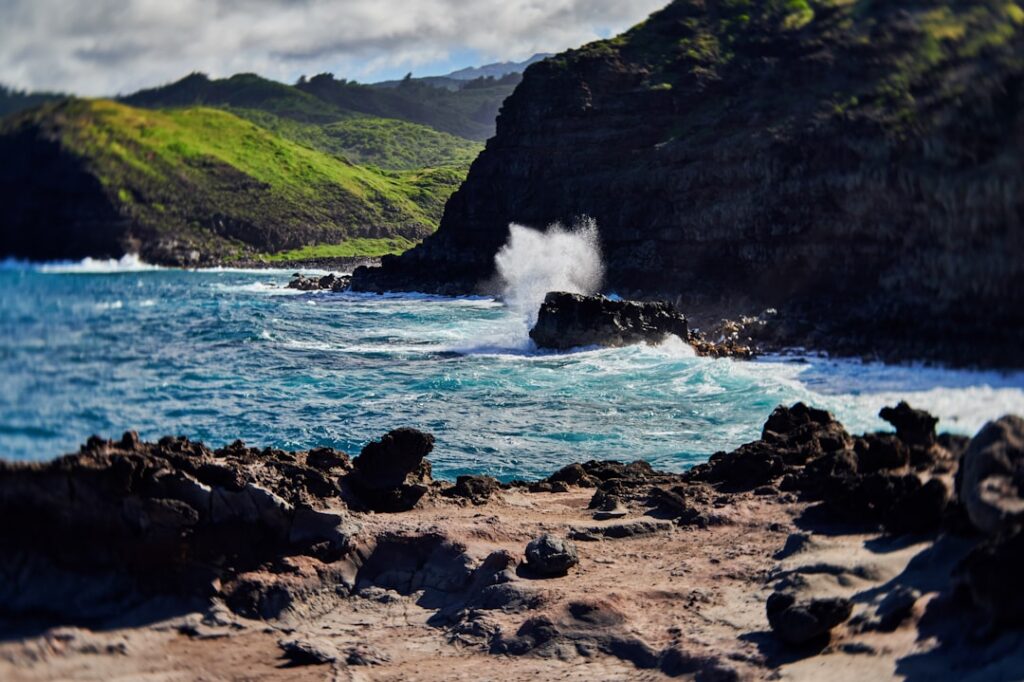 A landscape image of an ocean wave crashing along the rocky Maui coastline near the Nakalele Blowhole on a bright sunny day with lava rock in the foreground and lush green landscape in the background. Captured with a Lensbaby Velvet lens to create an ethereal, aesthetic vignette to the photograph th — Federal Aid Deadlines Approaching for March Kona Low Storm Damage | Maui Tom's News