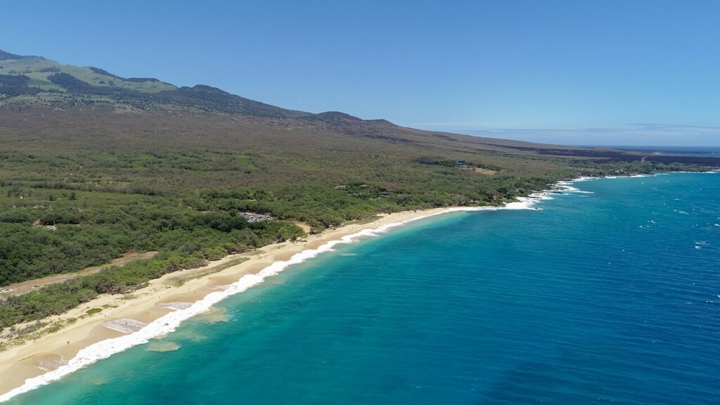 Aerial view of a tropical coastline with turquoise water, white-sand beach, and forested hills in the distance.
