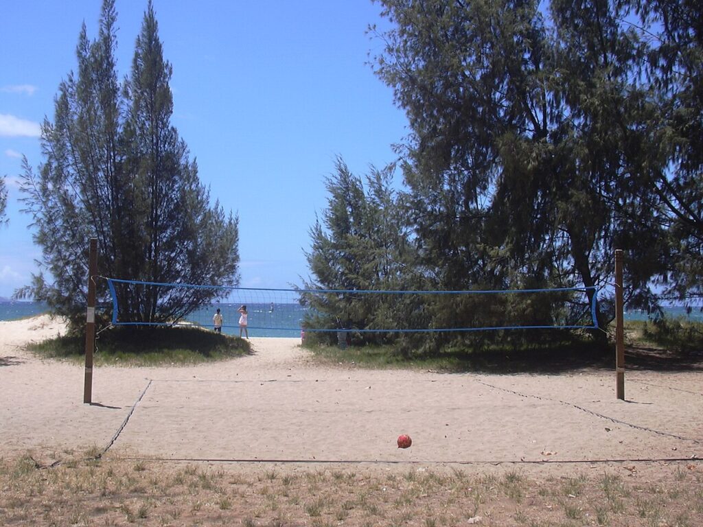 Beach volleyball court on a sandy beach with a blue net, ocean in the background and a few people near the shore.
