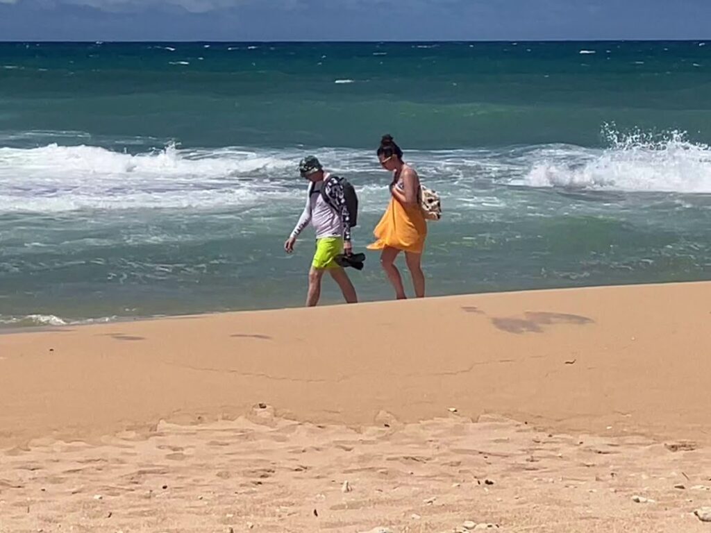 Two people walk along a sandy beach near the surf, one in a white shirt and neon shorts with a backpack, the other in an orange dress carrying a bag.