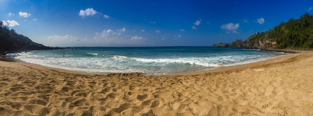 Panoramic beach scene with golden sand, blue ocean, small waves, and rocky headlands on both sides under a clear sky. A few swimmers are in the water.