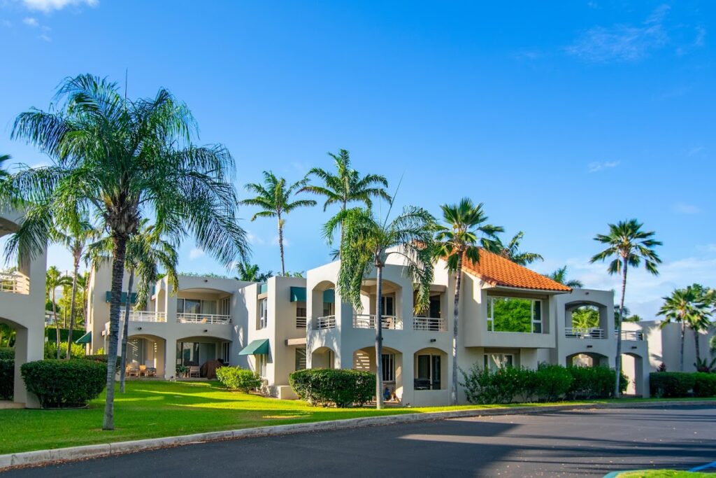 Two-story tropical resort buildings with arches, balconies, and palm trees under a clear blue sky.