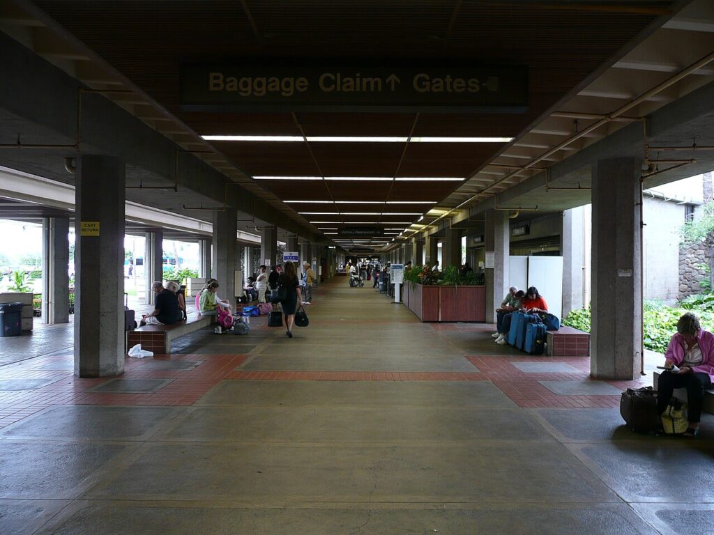 Long indoor terminal corridor with a 'Baggage Claim' sign overhead and people sitting with suitcases along the sides of the walkway.