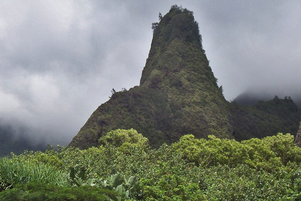 Cone-shaped, forest-covered mountain peak rising above dense tropical vegetation under a cloudy sky.