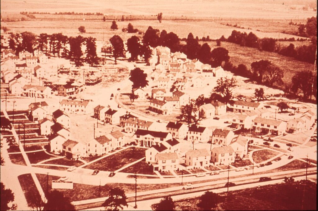 Aerial view of a small town with clustered houses, tree-lined streets, and open fields in the distance.