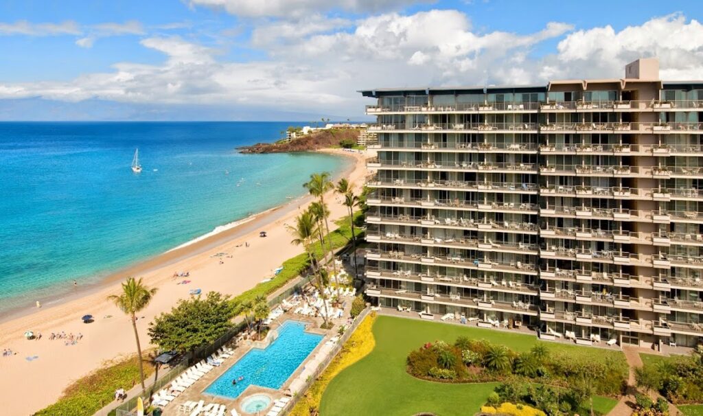 High-rise beachfront hotel with rows of balconies beside a sandy beach and turquoise sea, plus a rectangular pool in the foreground.