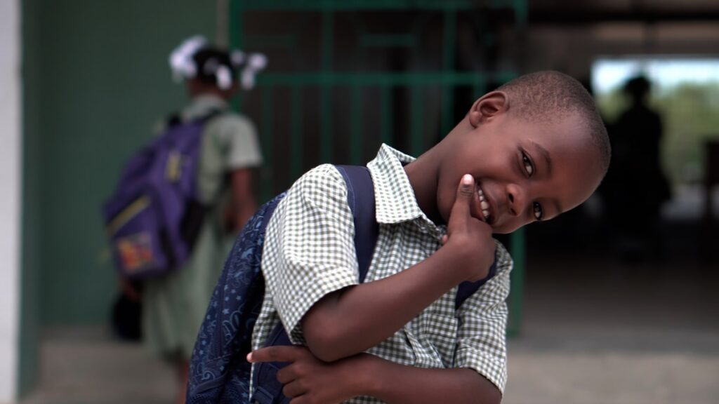 Smiling boy in a checkered shirt with a backpack tilting his head playfully at the camera in a school setting.