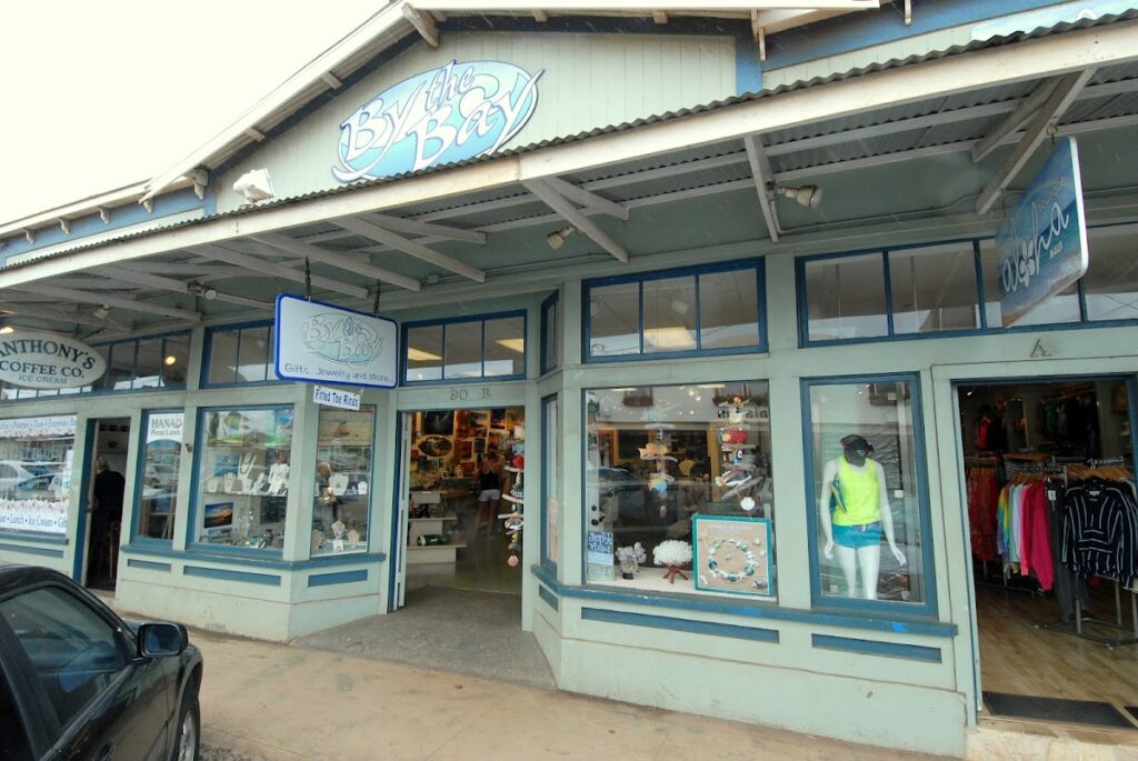 Coastal-style storefront 'By the Bay' with blue trim, displaying clothing and accessories in the windows.