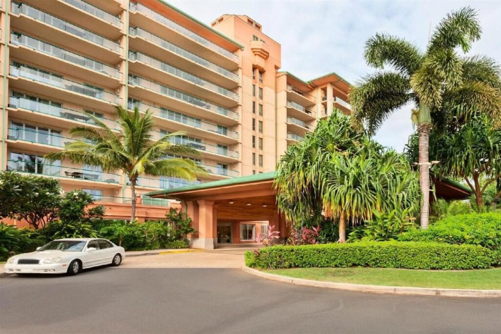 Hotel with multiple balconies, palm trees, and a circular driveway; a white car parked near the entrance.