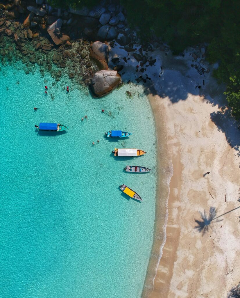 Aerial view of a tropical beach with turquoise water, several colorful boats anchored near a rocky shoreline and white sand, with palm tree shadows overhead.