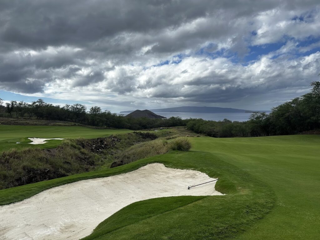 Coastal golf course with a green fairway, a large white sand bunker in the foreground, and the sea and hills under a cloudy sky.