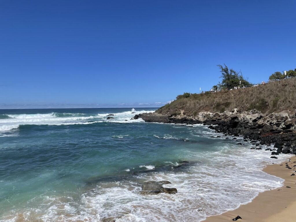 Clear blue sky over a rocky coastline; turquoise waves crash onto a sandy beach.