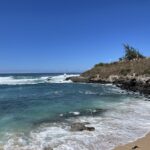 Potential Tropical System Could Threaten Maui by Next Weekend - Maui Tom's Vacation Rental Cleaning & Maintenance Clear blue sky over a rocky coastline; turquoise waves crash onto a sandy beach.