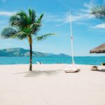 Three Years After Lahaina Fire, Commercial Rebuilding Lags Far Behind - Maui Tom's Vacation Rental Cleaning & Maintenance Sunny beach with palm trees, a small sailboat on the sand, and a thatched parasol by the turquoise water.