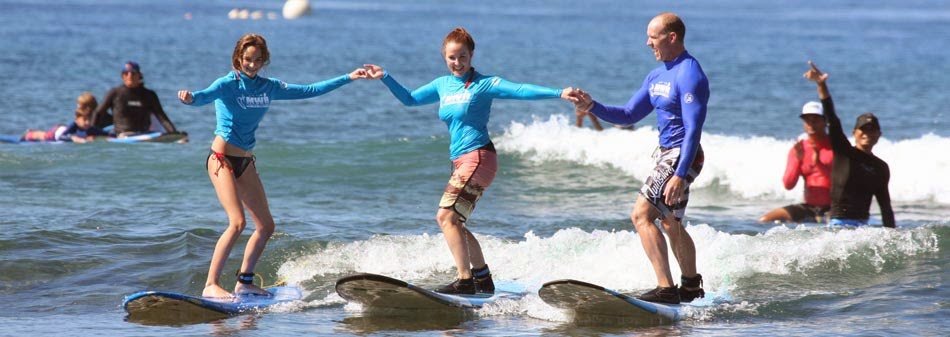 Two girls on surfboards in blue tops balance on small waves while a male instructor supports them nearby during a beach surf lesson.