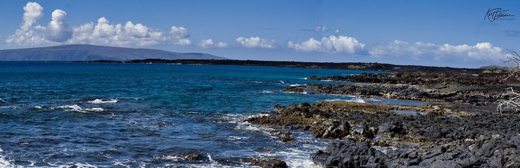 Coastal rocky shoreline with dark lava rocks, turquoise water, and a blue sky with white clouds.