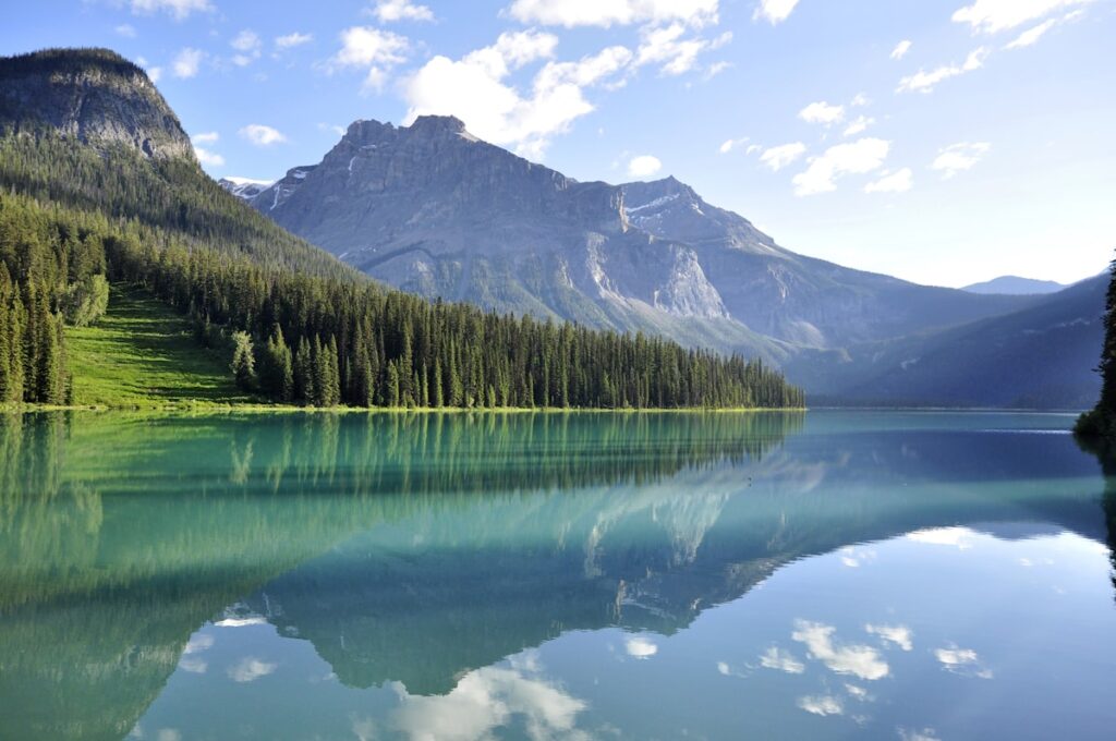 Alpine lake with evergreen forest along the shore, reflecting tall mountains under a blue sky with scattered clouds.