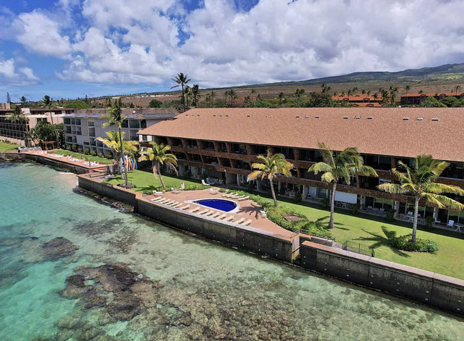 Aerial view of a tropical resort: brown-roofed buildings, palm trees, and a circular blue pool along the waterfront with turquoise water beyond the seawall.