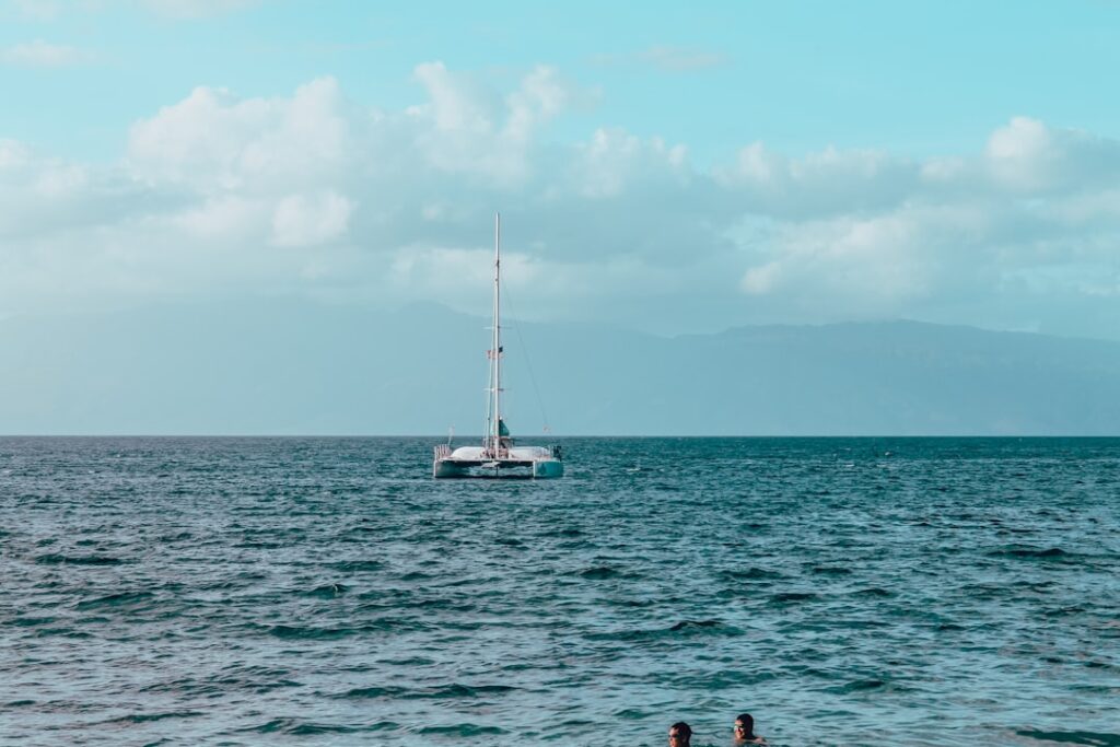 Sailboat on blue open water with two swimmers in the foreground and a cloudy sky on the horizon.