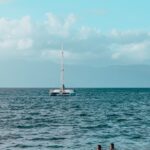 Sailboat on blue open water with two swimmers in the foreground and a cloudy sky on the horizon.